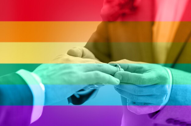 gente, homosexualidad, matrimonio entre personas del mismo sexo y concepto de amor - cierre de las manos de una pareja gay masculina feliz poniendo el anillo de bodas sobre el fondo de las rayas de la bandera del arco iris