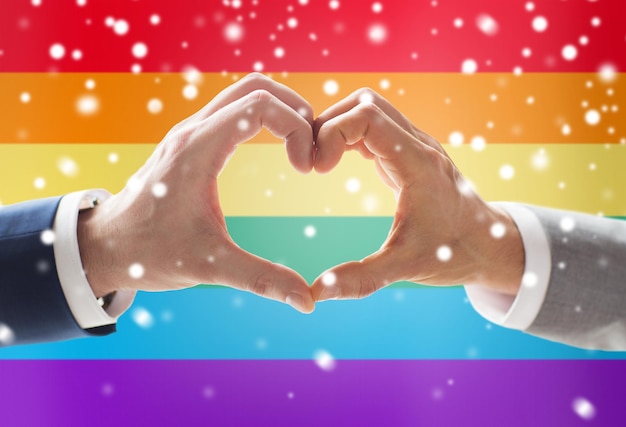 gente, homosexualidad, matrimonio entre personas del mismo sexo, gesto y concepto de amor - cierre de las manos de una pareja gay masculina feliz mostrando el signo de la mano del corazón sobre el fondo de la bandera del arco iris y el efecto de la nieve