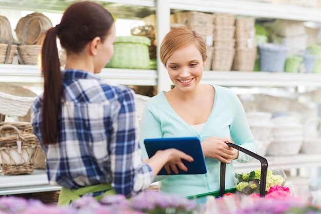 gente, jardinería, compras, venta y concepto de consumismo - jardinero feliz con tablet pc ayudando a la mujer a elegir flores en la floristería