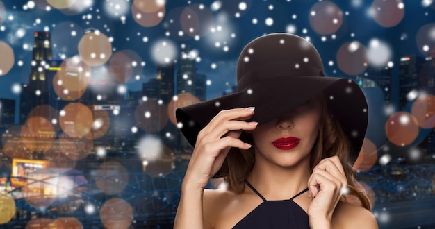 gente, lujo, navidad, vacaciones y concepto de moda - hermosa mujer con sombrero negro durante la noche luces de la ciudad de singapur fondo y nieve