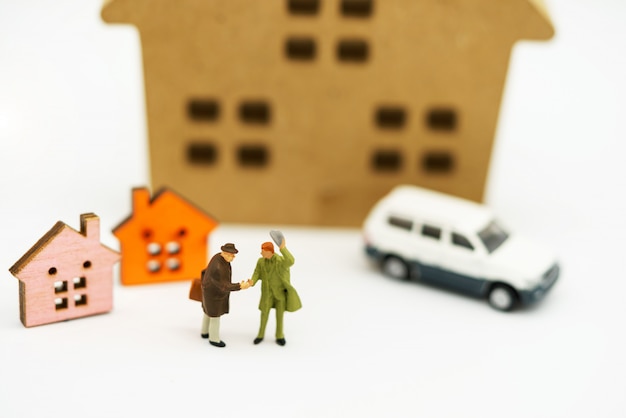 Gente en miniatura: apretón de manos de hombres de negocios frente a un bloque de madera con casa y automóvil.