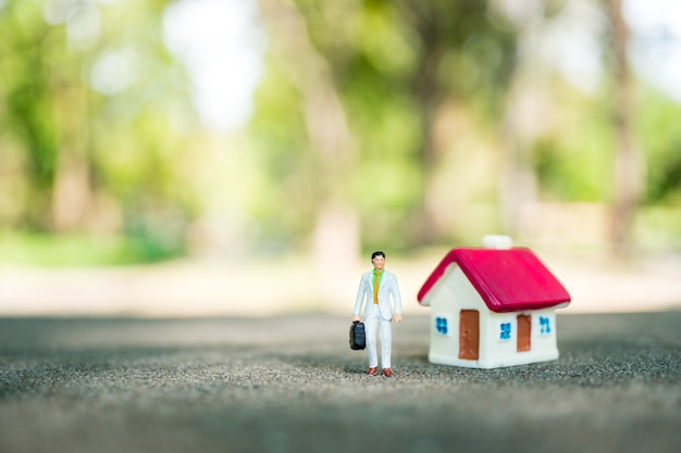 Gente miniatura, hombre de negocios que se coloca con la mini casa en fondo verde de la naturaleza usando como negocio,