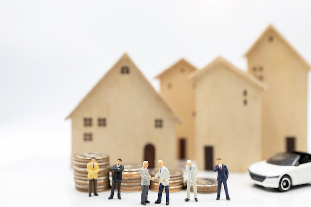 Gente en miniatura: hombres de negocios dándose la mano sobre la pila de monedas con casa y coche. Concepto de inversión en vivienda y vehículos.