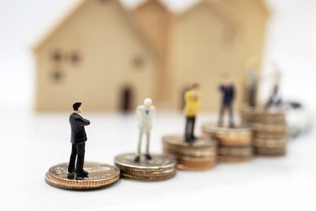 Gente en miniatura: hombres de negocios dándose la mano sobre la pila de monedas con casa y coche. Concepto de inversión en vivienda y vehículos.