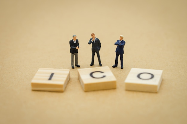 Gente en miniatura, hombres de negocios de pie con la palabra de madera ICO (Oferta inicial de monedas)