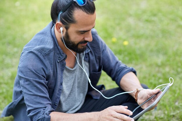 gente, música, tecnología, ocio y estilo de vida - feliz joven hipster con auriculares y tablet pc sentado en el césped