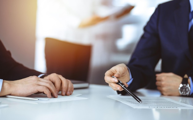 Gente de negocios discutiendo un contrato en una reunión o negociación, sentados juntos en el escritorio y una mujer desconocida en el fondo, cerca. Grupo de hombres de negocios desconocidos en una oficina moderna y soleada.