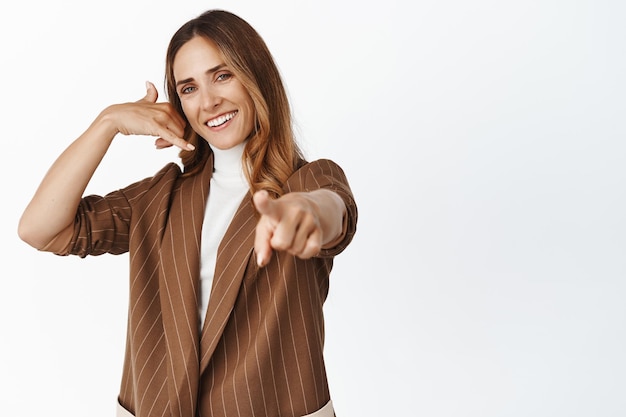 Gente de negocios Retrato de una mujer corporativa que muestra un gesto de llamada telefónica y señala con el dedo a la publicidad de la cámara de pie sobre fondo blanco