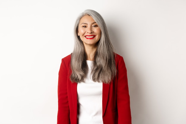 Gente de negocios. Senior mujer asiática sonriente con lápiz labial rojo y chaqueta mirando feliz, de pie sobre fondo blanco.