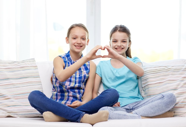 gente, niños, amigos y concepto de amistad - niñas felices sentadas en el sofá y mostrando el signo de la mano en forma de corazón en casa