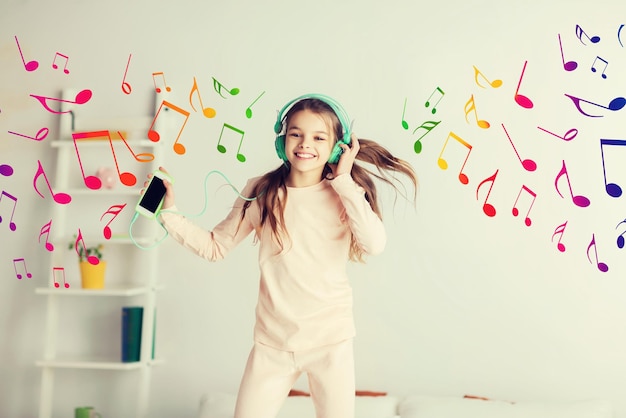 gente, niños, fiesta de pijamas y concepto de tecnología - chica sonriente feliz con auriculares saltando en la cama con smartphone y escuchando música en casa