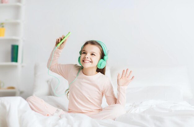 gente, niños, fiesta de pijamas y concepto de tecnología - chica sonriente feliz con auriculares sentada en la cama con smartphone y escuchando música en casa