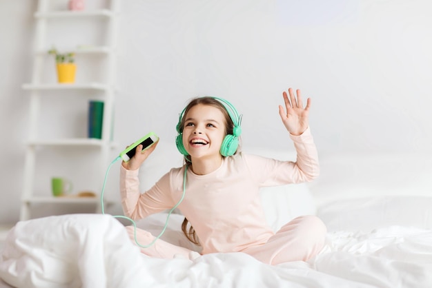 gente, niños, fiesta de pijamas y concepto de tecnología - chica sonriente feliz con auriculares sentada en la cama con smartphone y escuchando música en casa