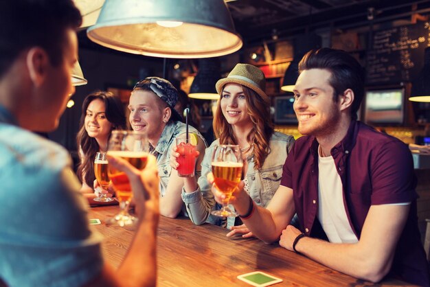 gente, ocio, amistad y concepto de comunicación - grupo de amigos sonrientes felices bebiendo cerveza y cócteles hablando en el bar o pub