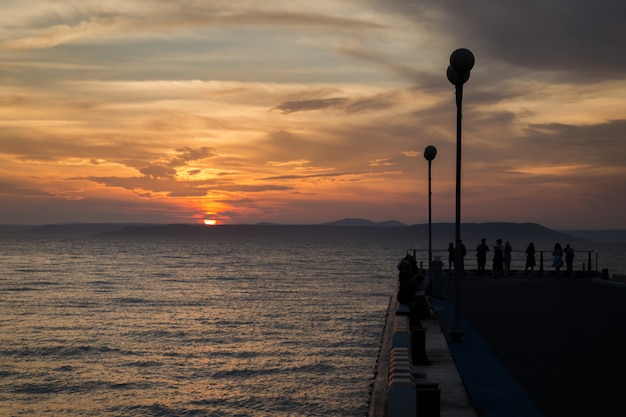 Gente de pie en el muelle y viendo colorido atardecer nublado