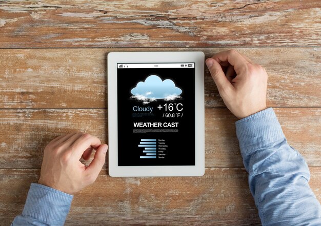 Foto gente, previsión meteorológica y concepto de tecnología - cierre de manos masculinas con el tiempo emitido en la pantalla de la computadora de tablet pc en la mesa de madera