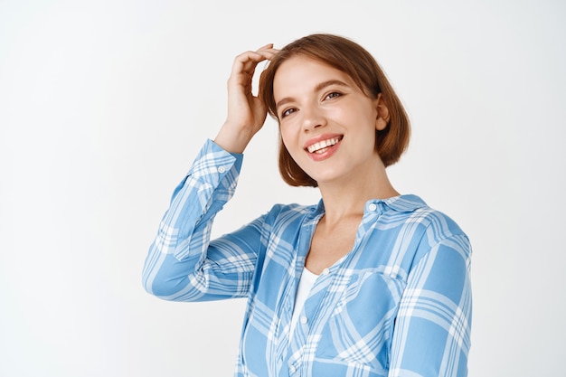 Gente real. Hermosa mujer caucásica sonriendo con dientes blancos, tocando el cabello y luciendo feliz. Chica joven con rostro bastante natural, arreglando el corte de pelo, de pie en la pared blanca