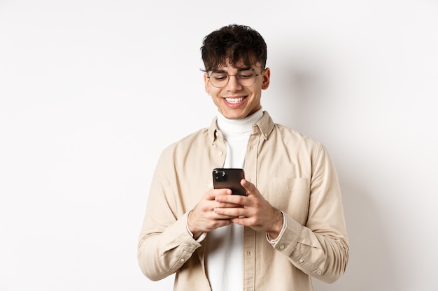 Gente real. Hombre joven natural leyendo el mensaje en el teléfono móvil, sonriendo y mirando la pantalla del teléfono inteligente, de pie en la pared blanca.