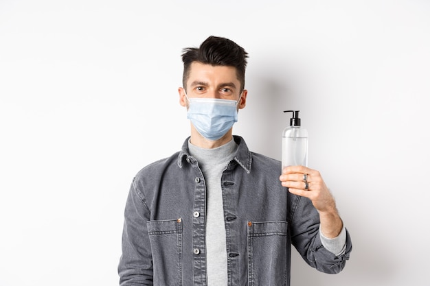 Gente sana y concepto de covid-19. Hombre joven con máscara médica usa medidas preventivas contra el coronavirus, mostrando una botella de desinfectante para manos, de pie contra el fondo blanco.