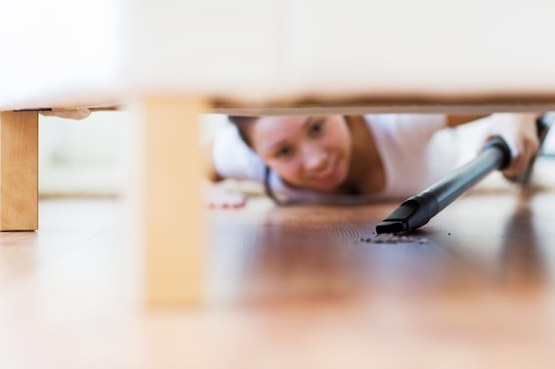 gente, tareas domésticas y concepto de limpieza - cerca de una mujer feliz con aspiradora limpiando el piso debajo del sofá en casa