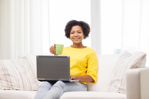 gente, tecnología y concepto de ocio - feliz joven afroamericana sentada en un sofá con ordenador portátil y bebiendo té de una taza en casa