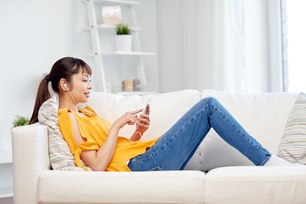 gente, tecnología y concepto de ocio - feliz joven asiática sentada en un sofá con smartphone y auriculares escuchando música en casa