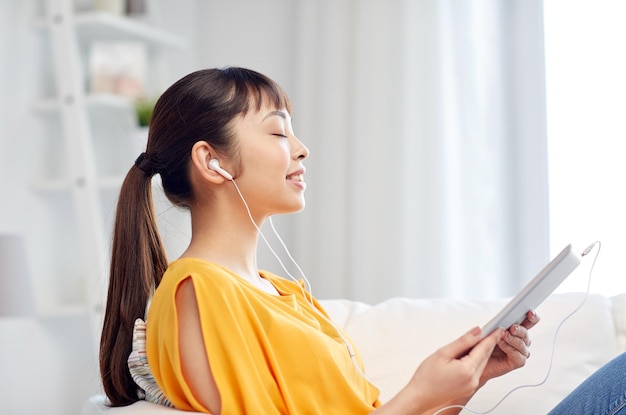 gente, tecnología y concepto de ocio - feliz joven asiática sentada en un sofá con tablet pc y auriculares escuchando música en casa