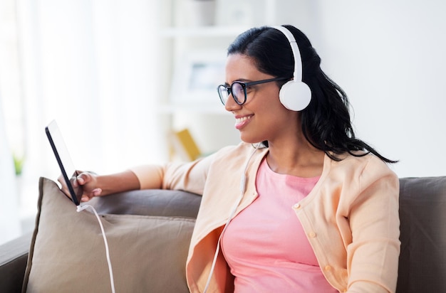 Gente, tecnología y concepto de ocio: una joven feliz sentada en un sofá con una tableta y auriculares escuchando música en casa