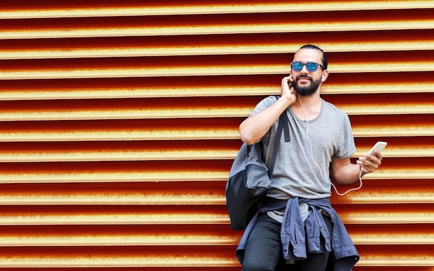 Foto gente tecnología viajes y turismo hombre con auriculares teléfono inteligente y bolsa en la calle de la ciudad y escuchar música sobre el fondo de la pared roja ribada