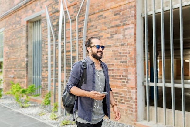 gente, tecnología, viajes y turismo - hombre con auriculares, teléfono inteligente y bolso caminando por la calle de la ciudad y escuchando música