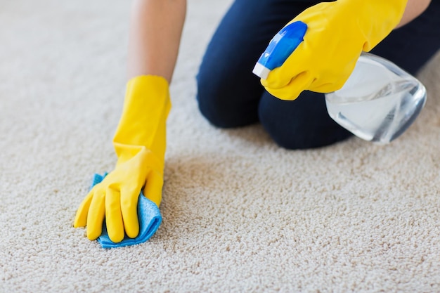 gente, trabajo doméstico y concepto de limpieza - cierre de mujer con guantes de goma con tela y alfombra de limpieza con spray detergente en casa
