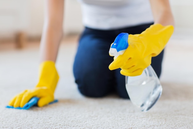 gente, trabajo doméstico y concepto de limpieza - cierre de mujer con guantes de goma con tela y alfombra de limpieza con spray detergente en casa