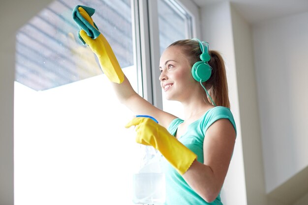 gente, trabajo doméstico y concepto de limpieza - mujer feliz con auriculares escuchando música y limpiando ventanas con limpiador en casa