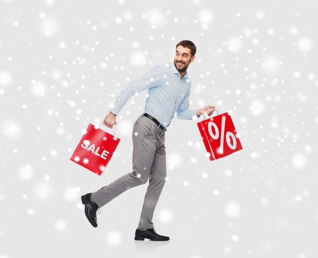 gente, venta, navidad, invierno y concepto de vacaciones - hombre sonriente sosteniendo bolsas de compras rojas con signo de porcentaje sobre fondo de nieve