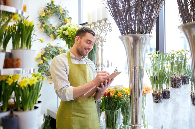 gente, venta, venta al por menor, negocios y concepto de floristería - feliz florista sonriente con portapapeles escribiendo y haciendo pedidos de notas en la floristería