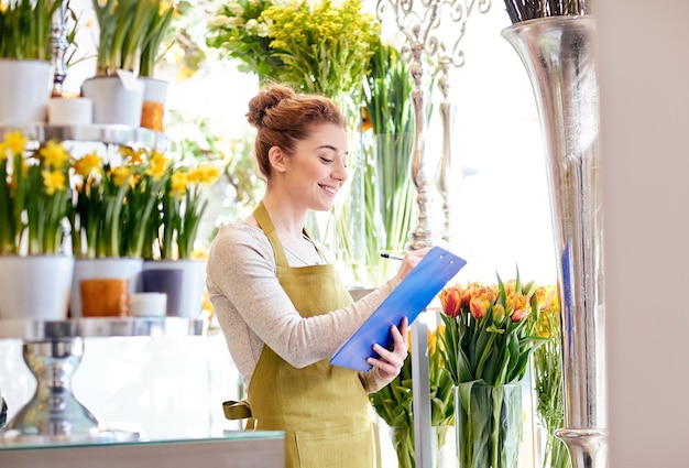 gente, venta, venta al por menor, negocios y concepto de floristería - mujer florista sonriente feliz con portapapeles escribiendo y haciendo pedidos de notas en la floristería