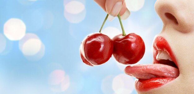 Foto gente, verano, concepto sexual y erótico - boca de mujer con labios y lengua comiendo cereza roja madura sobre fondo de luces azules