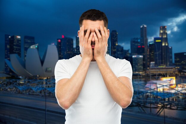 gente, viajes, turismo, emociones y concepto de estrés - hombre con camiseta blanca cubriendo su rostro con las manos sobre el fondo de la ciudad de singapur por la noche