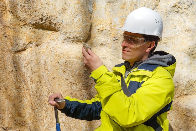 Geólogo masculino en casco y gafas protectoras examina una muestra del mineral al aire libre contra una roca