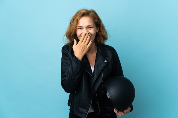 Georgianisches Mädchen, das einen Motorradhelm lokalisiert auf der blauen Wand glücklich und lächelnd hält Mund mit Hand hält