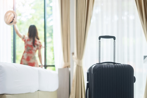 Gepäck im modernen Hotelzimmer mit der glücklichen jungen erwachsenen Frau, die fast Fenster entspannt, asiatische Touristin, die zur schönen Naturansicht schaut. Zeit zu reisen, Entspannung, Reise, Reise und Urlaubskonzepte