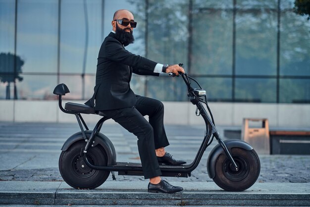 Gepflegter moderner Geschäftsmann mit Sonnenbrille fährt Elektroroller zu seinem Büro.