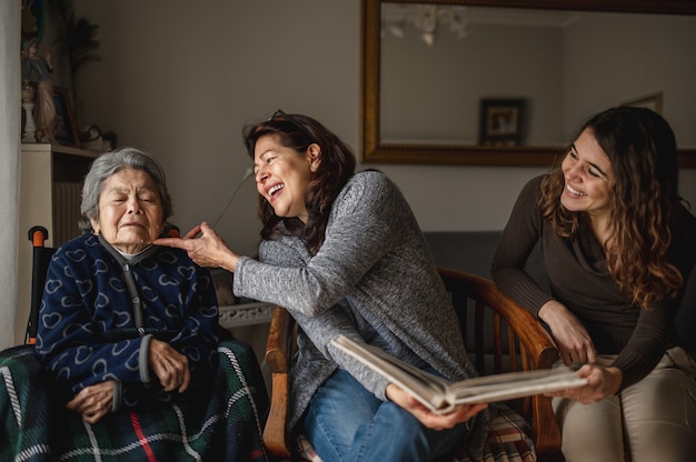 Geração de mulheres com a velha avó doente sentada na cadeira de rodas e sorrindo, filha e neta, olhando um álbum de fotos.