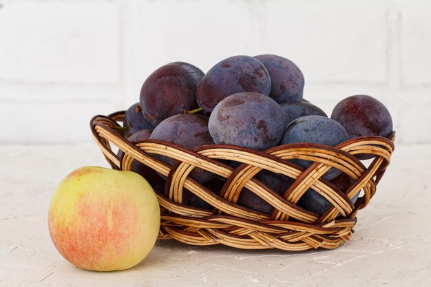 Gerade reife Pflaumen in einem Weidenkorb gepflückt und ein Apfel auf dem weißen strukturierten Hintergrund