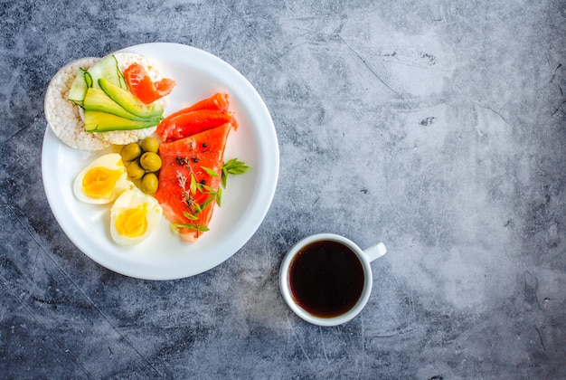 Geräucherter roter Fischlachs mit Oliven, Wachteleiern, Nüssen und knusprigen Reiskuchen mit Avocado auf grauem Hintergrund. Ansicht von oben. Rustikal. Produkte mit Omega-3 gut für gesundes Haar und gesunde Haut. Platz für Text.