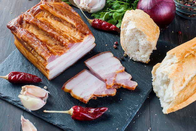 Geräucherter Speck mit Stangenbrot und Gemüse auf schwarzem Steinbrett