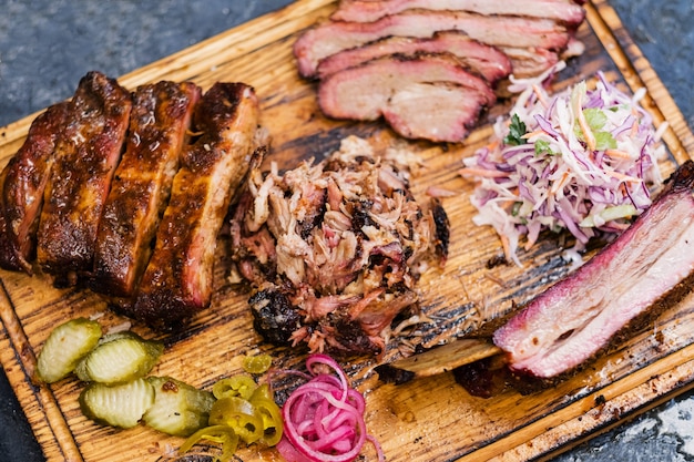 Geräuchertes Fleischsortiment auf Holzbrett. Draufsicht auf geschnittenes Rinderbrust, Pulled Pork, Rippchen, Krautsalat, Gurken.