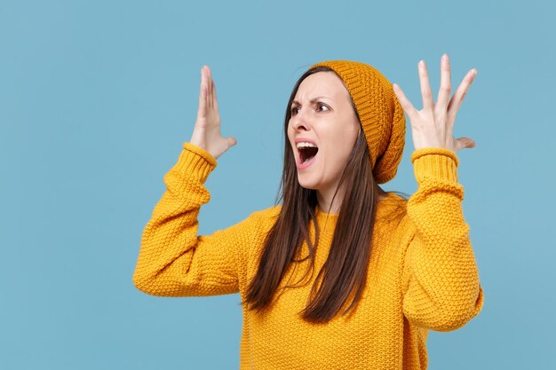 Gereizte junge brünette Frau Mädchen in gelbem Pullover und Hut posiert isoliert auf blauem Hintergrund Studioportrait. People-Lifestyle-Konzept. Mock-up-Kopienbereich. Zur Seite schauen und fluchend die Hände ausbreiten.