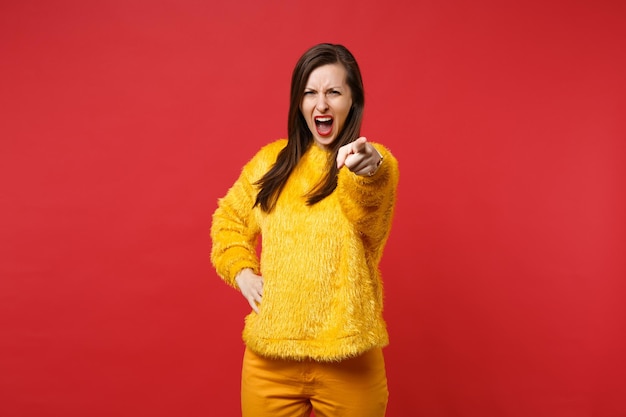 Gereizte junge Frau in gelbem Pelzpullover, die mit dem Zeigefinger auf die Kamera zeigt, schreit und flucht einzeln auf rotem Hintergrund im Studio. Menschen aufrichtige Emotionen Lifestyle-Konzept. Kopieren Sie Platz.