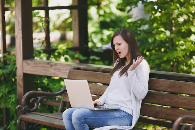 Gereizte und unzufriedene Geschäftsfrau in leichter Freizeitkleidung. Frau sitzt auf der Bank und arbeitet an einem modernen Laptop-PC im Stadtpark im Freien an der Natur. Mobiles Büro. Freiberufliches Geschäftskonzept